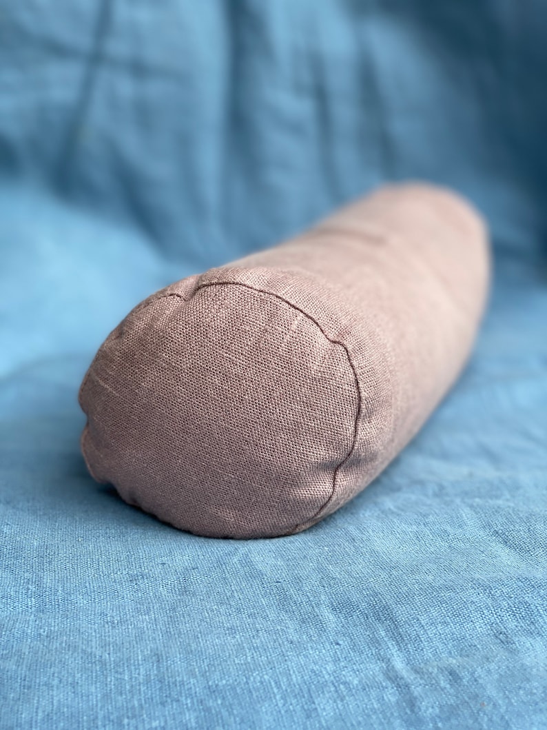 以下が含まれることがあります： 柔らかな薄い茶色の布で作られた円筒形の枕。枕は青い布の表面に置かれています。