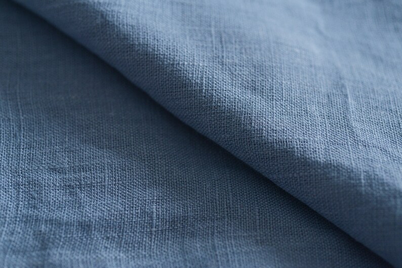 May include: Close-up of a blue linen fabric with a textured weave. The fabric is folded in half, showing the soft, natural texture.