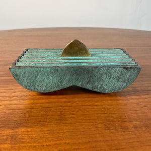 May include: A decorative box with a layered, rectangular design. The box has a teal patina finish and a brass triangle-shaped handle. The box is made of metal and sits on a wooden surface. The box is an art piece.