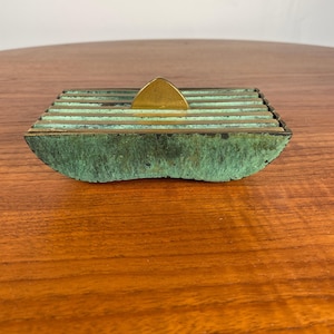 May include: A square, vintage ashtray with a verdigris finish and brass accents. The ashtray has a series of horizontal ridges and a triangular brass piece. The ashtray is made of metal and sits on a wooden surface.