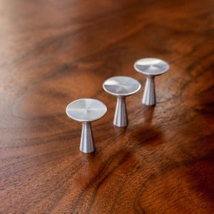 May include: Three silver metal cabinet knobs with a round top and a cylindrical base. The knobs are arranged in a row on a wooden surface.