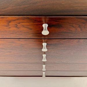 May include: A close-up of a wooden dresser with a rich, dark brown finish. The dresser has multiple drawers, each with a white, hourglass-shaped handle. The wood grain is visible, adding texture to the piece.
