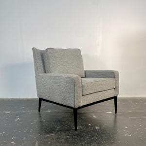 May include: A gray upholstered armchair with a boxy silhouette. The chair has a high back and armrests, with a black frame and legs. The fabric appears to be a textured weave. The chair is a modern style.