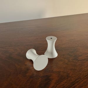 May include: Two light gray cabinet knobs with a rounded base and a cylindrical body. The knobs are made of a smooth material and are displayed on a dark wood surface.