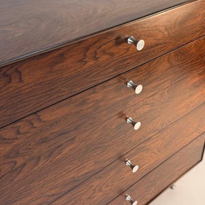 May include: A dark brown wooden dresser with five drawers. Each drawer has a silver, rounded knob. The wood grain is visible, and the finish is glossy. The dresser is a mid-century modern style.