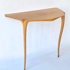 May include: A light brown, mid-century modern console table. The table has a unique, asymmetrical design with a half-moon shaped top and three slender, curved legs. The table is made of wood and has a smooth finish.