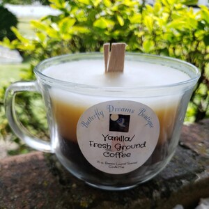 K&ouml;nnte beinhalten: Eine geschichtete Kerze in einer transparenten Glas-Tasse mit Henkel. Die Kerze hat drei Farbschichten: wei&szlig;, hellbraun und dunkelbraun. Das Etikett lautet "Vanilla/ Fresh Ground Coffee". Ein Holzdocht befindet sich in der Mitte.