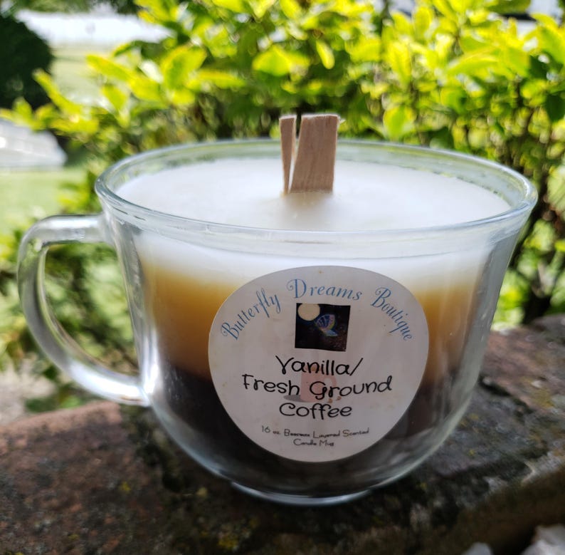K&ouml;nnte beinhalten: Eine geschichtete Kerze in einer transparenten Glas-Tasse mit Henkel. Die Kerze hat drei Farbschichten: wei&szlig;, hellbraun und dunkelbraun. Das Etikett lautet "Vanilla/ Fresh Ground Coffee". Ein Holzdocht befindet sich in der Mitte.