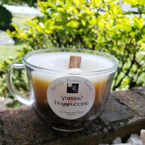K&ouml;nnte beinhalten: Eine Kerze in Form einer Kaffeetasse aus klarem Glas mit einem geschichteten Aussehen. Die Kerze hat eine wei&szlig;e Oberseite, eine beige Mitte und eine dunkelbraune Basis. Sie hat einen Holzdocht und ein Etikett mit der Aufschrift "Vanilla/Frappuccino".