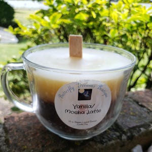K&ouml;nnte beinhalten: Eine Vanilla Mocha Latte Duftkerze in einer transparenten Glas-Tasse. Die Kerze hat ein geschichtetes Aussehen mit einer hellen cremefarbenen Oberseite, einer tanfarbenen Mitte und einer dunkelbraunen Basis. Ein Holzdocht befindet sich in der Mitte. Das Etikett lautet "Butterfly Dreams Boutique".
