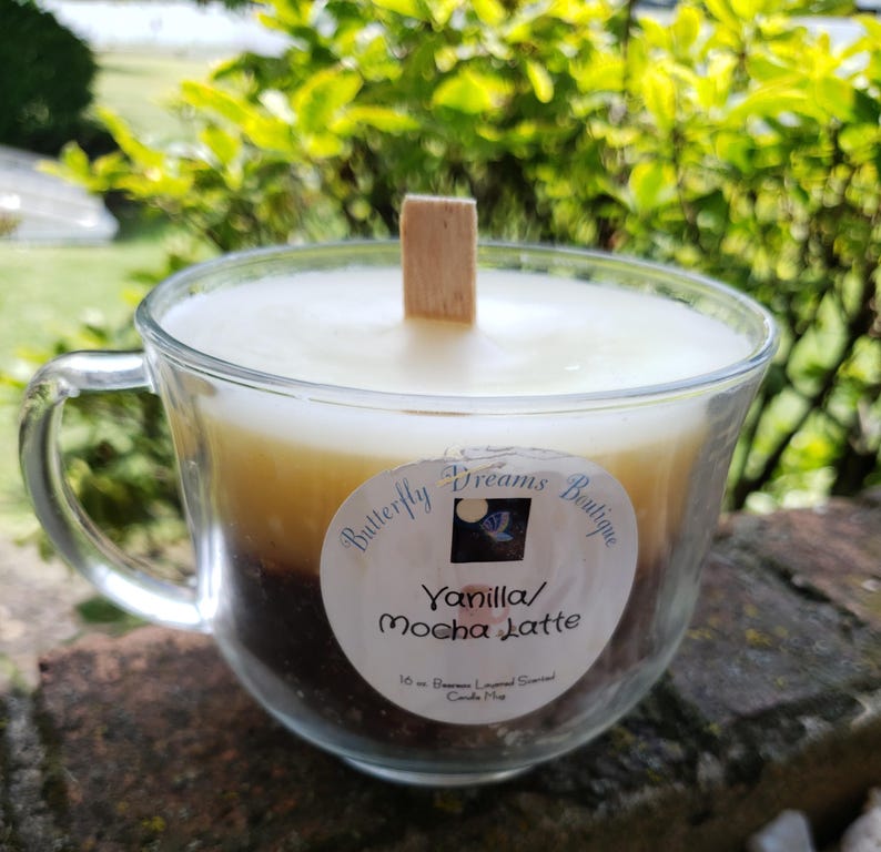 K&ouml;nnte beinhalten: Eine Vanilla Mocha Latte Duftkerze in einer transparenten Glas-Tasse. Die Kerze hat ein geschichtetes Aussehen mit einer hellen cremefarbenen Oberseite, einer tanfarbenen Mitte und einer dunkelbraunen Basis. Ein Holzdocht befindet sich in der Mitte. Das Etikett lautet "Butterfly Dreams Boutique".