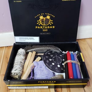 May include: An open black cigar box with the text "PARTAGAS" and "CLASICO 5 1/4X54". Inside are sage, feathers, candles, a small bag, and a moon phase chart. A purple Sharpie marker is in the foreground.