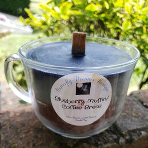 K&ouml;nnte beinhalten: Eine Kerze in Form einer Teetasse aus klarem Glas mit einer dunkelblauen Wachsoberfl&auml;che und einem Holzdocht. Die Kerze hat ein Etikett mit der Aufschrift "Butterfly Dreams Boutique" und "Blueberry Muffin/Coffee Break". Die Kerze steht auf einer Steinoberfl&auml;che.