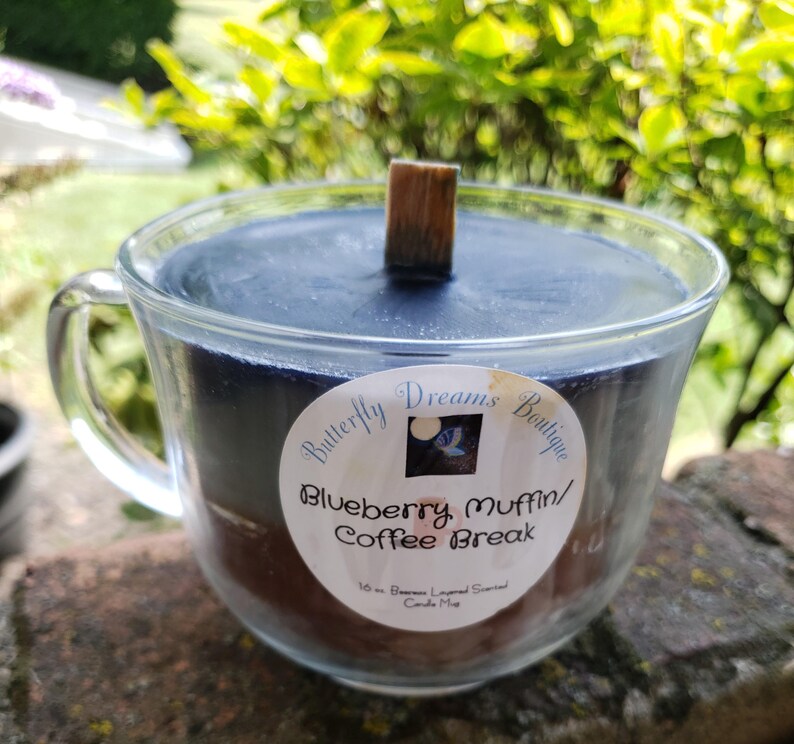 K&ouml;nnte beinhalten: Eine Kerze in Form einer Teetasse aus klarem Glas mit einer dunkelblauen Wachsoberfl&auml;che und einem Holzdocht. Die Kerze hat ein Etikett mit der Aufschrift "Butterfly Dreams Boutique" und "Blueberry Muffin/Coffee Break". Die Kerze steht auf einer Steinoberfl&auml;che.