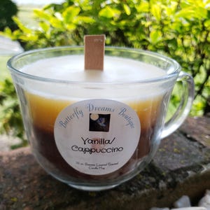 K&ouml;nnte beinhalten: Geschichtete Vanilla/Cappuccino Duftkerze in einer transparenten Glas-Tasse. Die Kerze hat einen Holzdocht und ein Etikett mit der Aufschrift "Butterfly Dreams Boutique". Die Kerze ist 283 Gramm.