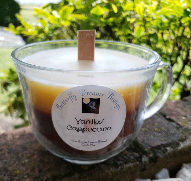 K&ouml;nnte beinhalten: Geschichtete Vanilla/Cappuccino Duftkerze in einer transparenten Glas-Tasse. Die Kerze hat einen Holzdocht und ein Etikett mit der Aufschrift "Butterfly Dreams Boutique". Die Kerze ist 283 Gramm.