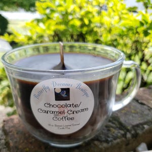 K&ouml;nnte beinhalten: Eine Kerze in Form einer Kaffeetasse aus klarem Glas, gef&uuml;llt mit dunkelbraunem Wachs. Die Kerze hat einen Holzdocht und ein Etikett mit der Aufschrift "Butterfly Dreams Boutique Chocolate/Caramel Cream Coffee". Die Kerze ist 454 Gramm.