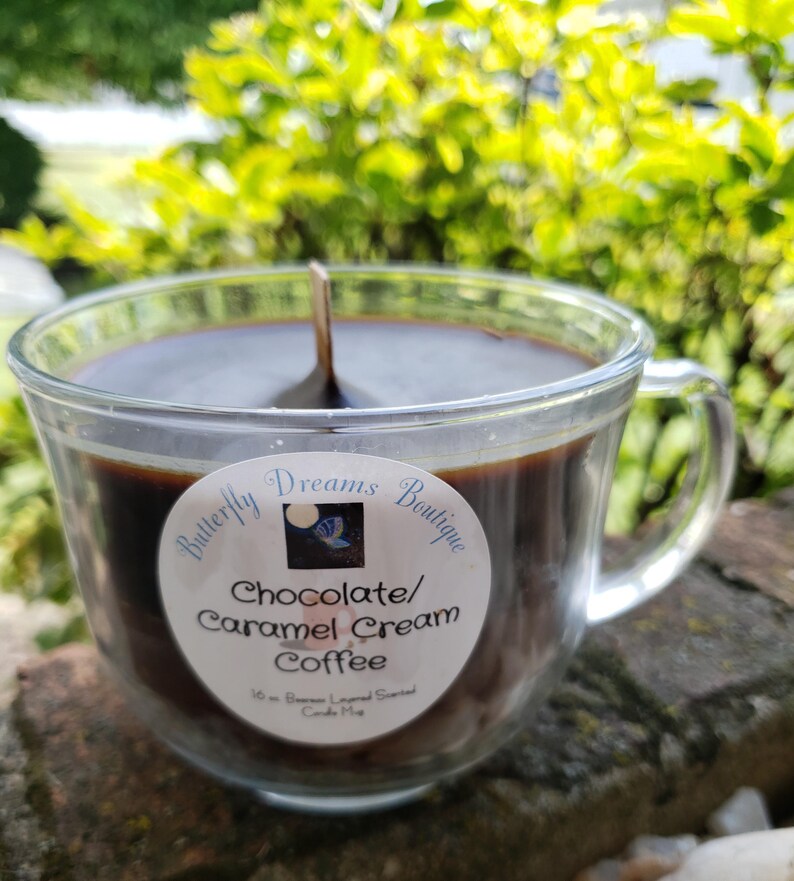 K&ouml;nnte beinhalten: Eine Kerze in Form einer Kaffeetasse aus klarem Glas, gef&uuml;llt mit dunkelbraunem Wachs. Die Kerze hat einen Holzdocht und ein Etikett mit der Aufschrift "Butterfly Dreams Boutique Chocolate/Caramel Cream Coffee". Die Kerze ist 454 Gramm.