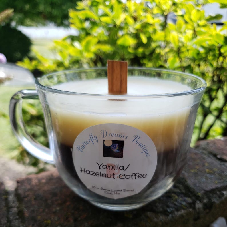 K&ouml;nnte beinhalten: Eine Kerze in einem transparenten Glas-Kaffeebecher mit einem Holzdocht. Die Kerze hat drei Schichten: wei&szlig;, beige und braun. Das Etikett lautet "Butterfly Dreams Boutique" und "Vanilla/Hazelnut Coffee".