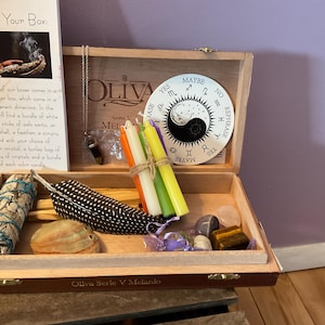 Op de afbeelding: Een open houten kist met diverse spirituele items. Binnenin bevinden zich saliebundels, kaarsen, kristallen, een veer en een decoratief yin-yang bord. Een kaart links leest "About Your Box".