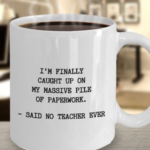 May include: White ceramic coffee mug with black text that reads "I'm finally caught up on my massive pile of paperwork. - Said no teacher ever."