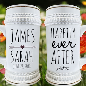 May include: Two white ceramic beer steins with black lettering. The left stein says "James & Sarah June 28, 2018" with an arrow and heart. The right stein says "Happily Ever After" with a small leaf design.