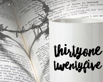 Könnte beinhalten: Eine weiße Keramik-Kaffeetasse mit schwarzem Text, der "thirtyone twentyfive" lautet. Die Tasse steht auf einem aufgeschlagenen Buch.