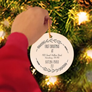 May include: A white round Christmas ornament with black text that reads "First Christmas at 1903 David Hollow Road, Harrisburg, PA 17112 Kaitlin & Mario 2021".