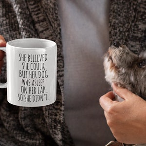 Op de afbeelding: Een witte koffiemok met zwarte tekst die luidt: "Ze geloofde dat ze kon, maar haar hond sliep op haar schoot... dus deed ze het niet."