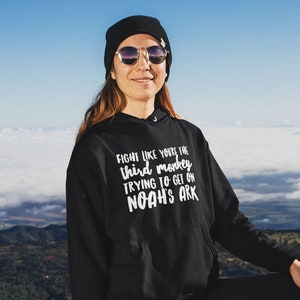 May include: A person wearing a black hoodie with the text "Fight like you're the third monkey trying to get on Noah's Ark" in white. The person is standing on a rocky mountaintop with a golden retriever dog.