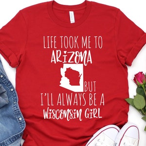 May include: A red t-shirt with white text that reads 'Life took me to Arizona but I'll always be a Wisconsin girl.' The shirt is laid out on a white surface with a pair of white sneakers, a denim jacket, and a bouquet of pink roses.