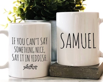 May include: Two white ceramic mugs. One mug has the text "IF YOU CAN'T SAY SOMETHING NICE, SAY IT IN YIDDISH." with a small leaf design. The other mug has the name "SAMUEL" printed on it.