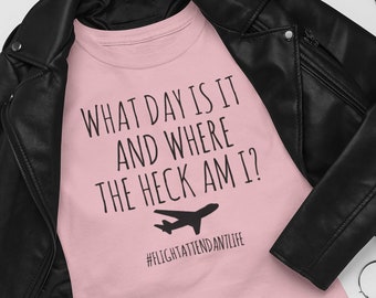 May include: Pink t-shirt with the text "What day is it and where the heck am I?" and a small airplane graphic. The text "#flightattendantlife" is below the graphic. The t-shirt is laid out on a white surface with a black leather jacket, black boots, lipstick, and a small black purse with silver studs.