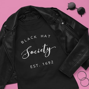 May include: Black t-shirt with the text "Black Hat Society Est. 1692" in white lettering. The shirt is laid out on a pink background with a black leather jacket, black boots, a black studded purse, lipstick, and earrings.