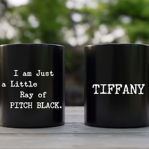 May include: Two black coffee mugs. One mug says "I am Just a Little Ray of PITCH BLACK." The other mug says "TIFFANY".