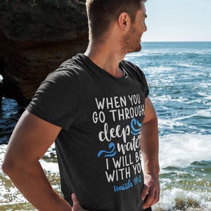 May include: Dark gray t-shirt with the text "WHEN YOU GO THROUGH deep water I WILL BE WITH YOU Isaiah 43:2" in white and blue. The shirt is worn by a person standing on a beach with the ocean in the background.