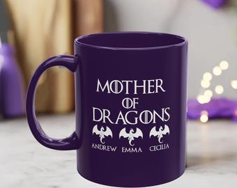 May include: A dark purple ceramic mug featuring the white text "MOTHER OF DRAGONS" with a sword icon replacing the letter O. Below the text are three white dragon silhouettes, each positioned above the names Andrew, Emma, and Cecilia.
