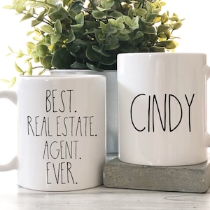 May include: Two white ceramic mugs with black text. One mug says "BEST. REAL ESTATE. AGENT. EVER." and the other mug says "CINDY".