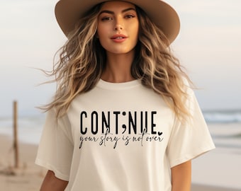 May include: A white t-shirt with the text "CONTINUE. your story is not over" printed in black. The shirt is being worn by a person with long blonde hair and a brown hat.