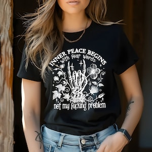 May include: Black t-shirt with a white graphic design featuring a skeleton hand making a rock and roll gesture with the text "Inner peace begins with four words: not my fucking problem".