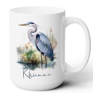 May include: White ceramic mug featuring a watercolor illustration of a Great Blue Heron standing in water with plants. The heron is depicted in shades of blue, gray, and black, with a yellow beak. The name "Rhianna" is written in cursive below the bird.