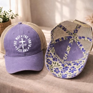 May include: A purple baseball cap with a mesh back and a floral pattern on the inside. The front of the cap features the text "THE WAY THE TRUTH THE LIFE" surrounding a cross and the word "JESUS".