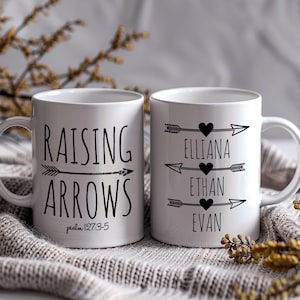 May include: Two white ceramic mugs with black text and arrows. One mug says "Raising Arrows" with the scripture "Psalm 127:3-5" below. The other mug has three arrows with hearts and the names "Elliana", "Ethan", and "Evan" written below.