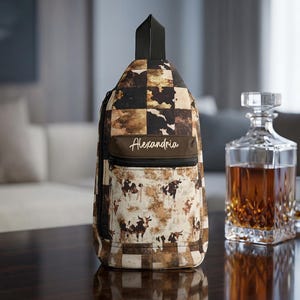 May include: A sling backpack featuring a brown and cream cow print pattern mixed with a checkered design. The bag includes a front zippered pocket with the name Alexandria printed in white script, a black top handle, and black zipper hardware.