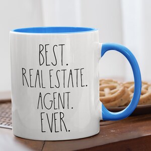 May include: White ceramic coffee mug with a blue rim and handle. The mug has black text that reads "BEST. REAL ESTATE. AGENT. EVER."