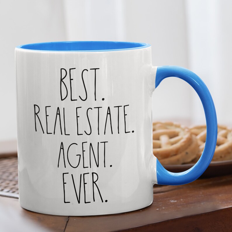 May include: White ceramic coffee mug with a blue rim and handle. The mug has black text that reads "BEST. REAL ESTATE. AGENT. EVER."