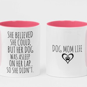 Op de afbeelding: Twee witte koffiemokken met roze accenten. Op de ene mok staat "She believed she could, but her dog was asleep on her lap... so she didn't." Op de andere mok staat "Dog Mom Life" met een hart en een pootprint.
