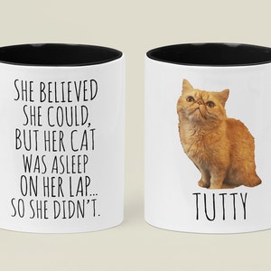 Puede incluir: Taza de cer&aacute;mica blanca con borde y asa negros. La taza tiene un texto negro que dice "SHE BELIEVED SHE COULD, BUT HER CAT WAS ASLEEP ON HER LAP... SO SHE DIDN'T." El otro lado de la taza tiene una imagen de un gato naranja con el nombre "TUTTY" escrito debajo.