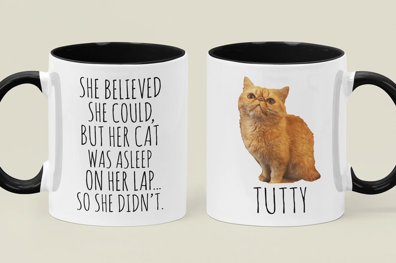 Puede incluir: Taza de cer&aacute;mica blanca con borde y asa negros. La taza tiene un texto negro que dice "SHE BELIEVED SHE COULD, BUT HER CAT WAS ASLEEP ON HER LAP... SO SHE DIDN'T." El otro lado de la taza tiene una imagen de un gato naranja con el nombre "TUTTY" escrito debajo.