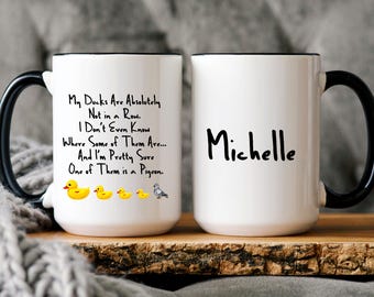 May include: Two white ceramic mugs with black handles. The mug on the left has a black rim and a humorous saying about ducks. The mug on the right has a black rim and the name "Michelle" written in black.